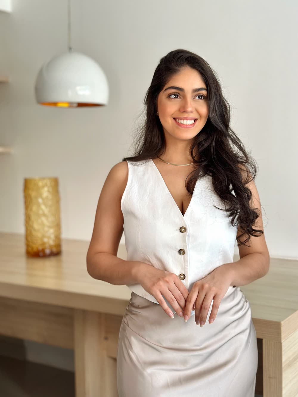 Daffine Benaion em pé, de colete branco, apoiada em uma bancada de madeira clara, sorrindo.